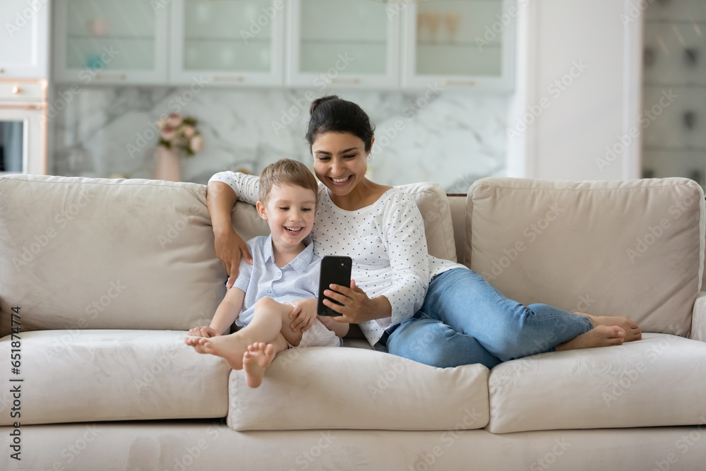 Happy Indian mother and Caucasian 7s son using smartphone together, relaxing sitting on cozy couch at home, smiling young mom with boy child looking at phone screen, watching cartoons, having fun
