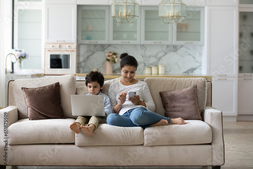 Smiling Indian mother and 5s boy using gadgets at home, sitting on couch together, young mom holding smartphone and little son spending leisure time with devices, addiction and overuse