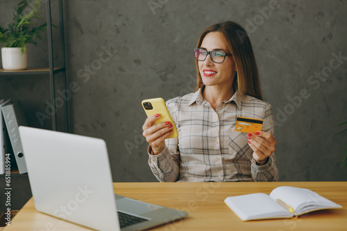 Young employee business woman wearing shirt casual clothes glasses sit work at office desk with pc laptop using mobile cell phone hold credit bank card shoppingonline. Achievement career job concept. photo