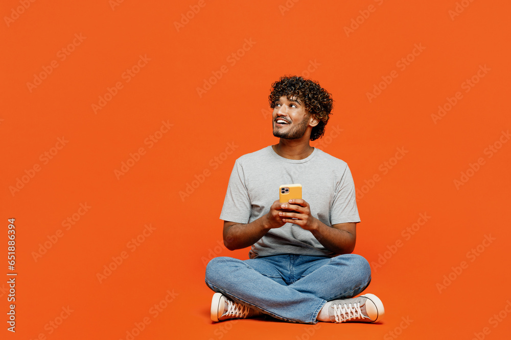 Full body young smiling happy Indian man he wears t-shirt casual clothes sit hold in hand use mobile cell phone look aside isolated on orange red color background studio portrait. Lifestyle concept. Full body young smiling happy Indian man he wears t-shirt casual clothes sit hold in hand use mobile cell phone look aside isolated on orange red color background studio portrait. Lifestyle concept.