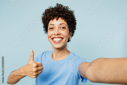 Close up young happy fun woman of African American ethnicity wear t-shirt casual clothes doing selfie shot pov on mobile cell phone show thumb up isolated on plain pastel light blue cyan background.
