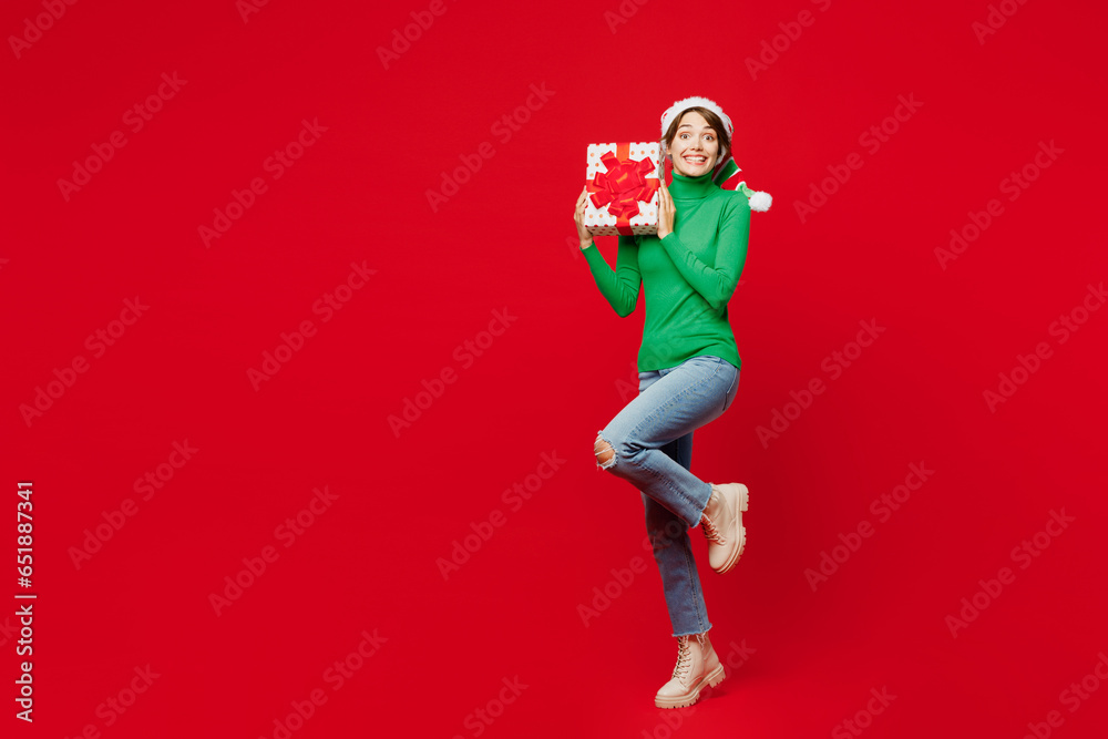 Full body overjoyed young woman wear green turtleneck Santa hat posing hold present box with gift ribbon bow isolated on plain red background Happy New Year 2024 celebration Christmas holiday concept Full body overjoyed young woman wear green turtleneck Santa hat posing hold present box with gift ribbon bow isolated on plain red background Happy New Year 2024 celebration Christmas holiday concept