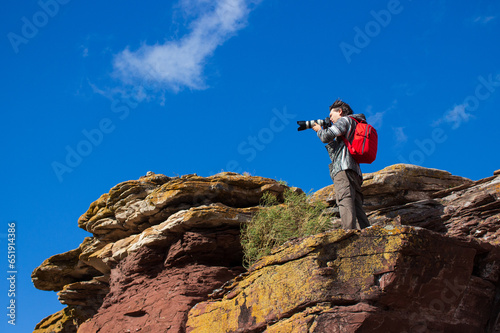 A male photographer stands on the edge of a cliff and takes pictures. A brunet with a red backpack at mountain against blue sky background. Shot of a man taking photos with his camera at travelling