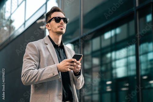 Serious frowning man wearing smart casual business clothes walking on the urban street and holding a mobile phone in his hands.