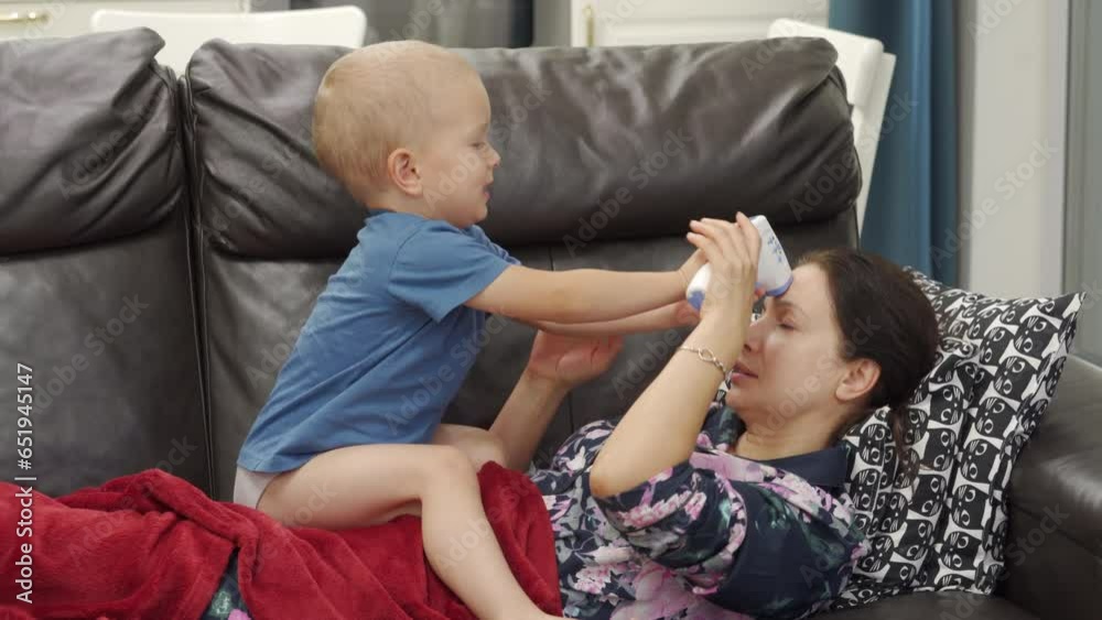 sick toddler child little boy holding infrared laser forehead thermometer playing with mother on sofa at home sick toddler child little boy holding infrared laser forehead thermometer playing with mother on sofa at home
