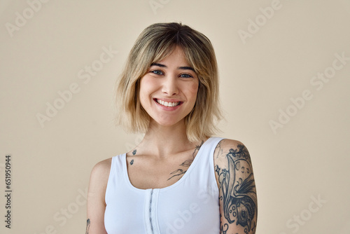 Young happy blond pretty smiling girl beauty female gen z model with short blonde hair beautiful face healthy skin and tattoo looking at camera wearing white top isolated at beige background. Portrait photo