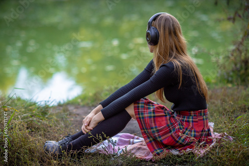 A long-haired schoolgirl with black socks, a black sweater, black shoes and a red skirt is sitting on the riverbank with headphones and listening to music. Soft focus