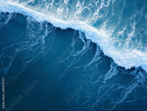 aerial perspective, the deep navy blue expanse of the ocean stretches out beneath, with white waves crashing and splashing against its surface.