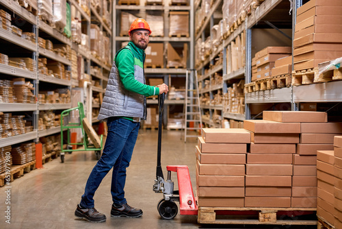 caucasian warehouse worker with manual pallet jack, working in storage room, transporting cardboard goods, boxes delivery. logistics and distribution concept. man in uniform look at camera smiling