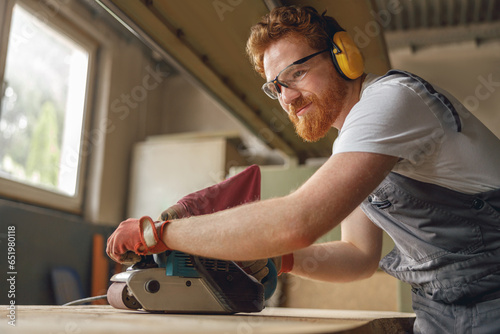 Bearded male wood worker using electric sander for wood. Carpentry workshop. High quality photo