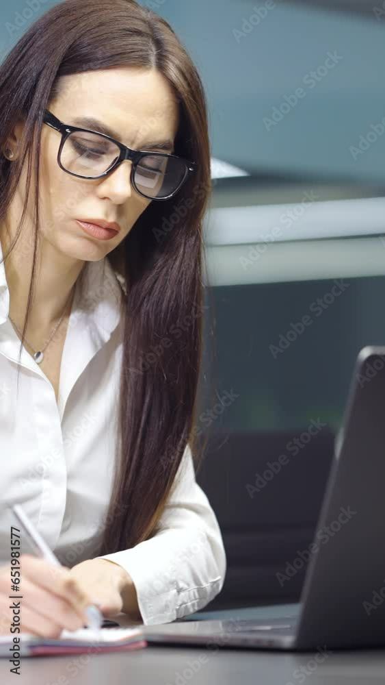 Vertical Screen: Businesswoman with long hair wearing white shirt and eyeglasses writing in notebook, scheduling meetings, laptop in front of her. Top manager at work. Concept of planning Vertical Screen: Businesswoman with long hair wearing white shirt and eyeglasses writing in notebook, scheduling meetings, laptop in front of her. Top manager at work. Concept of planning