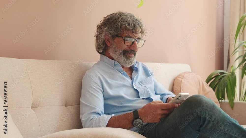 An elderly man sits on sofa in cozy living-room with phone in his hands An elderly man sits on sofa in cozy living-room with phone in his hands