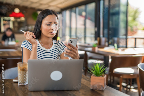One young woman sitting in cafe holding electronic cigarette and looking at mobile phone, modern lifestyle concept