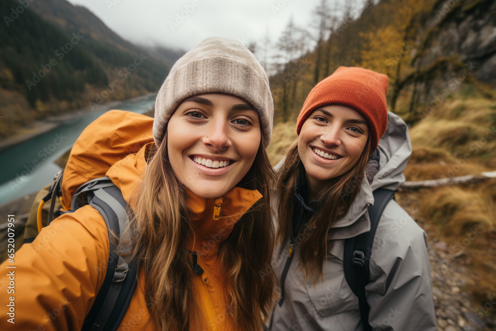 Two female friends taking selfies by a river during a hike along the mountain.