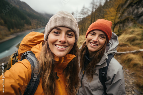 Two female friends taking selfies by a river during a hike along the mountain.