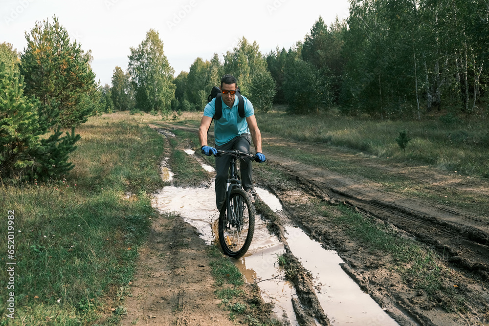 man wearing sports gloves and carrying a backpack rides a mountain bike through a puddle in a forest area.Active lifestyle