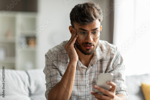 Shocked indian guy looking at cellphone screen, sitting on couch at home, holding phone and touching his head, reading bad news or message, copy space