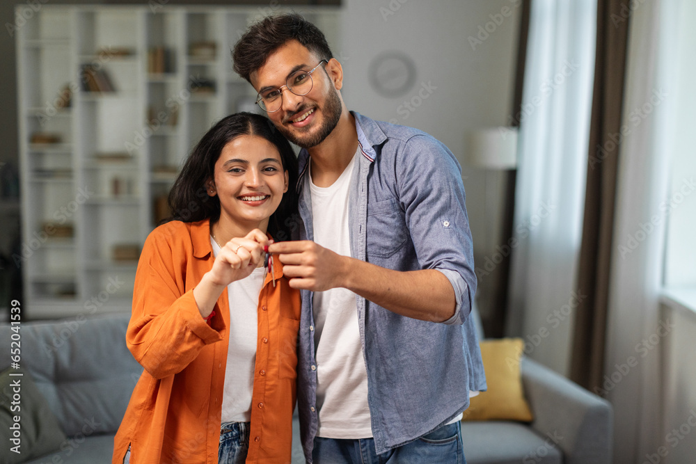 Excited young indian husband and wife showing keys from their new apartment, enjoy buying own home, standing in room interior, copy space. Mortgage real estate concept - obrazy, fototapety, plakaty 