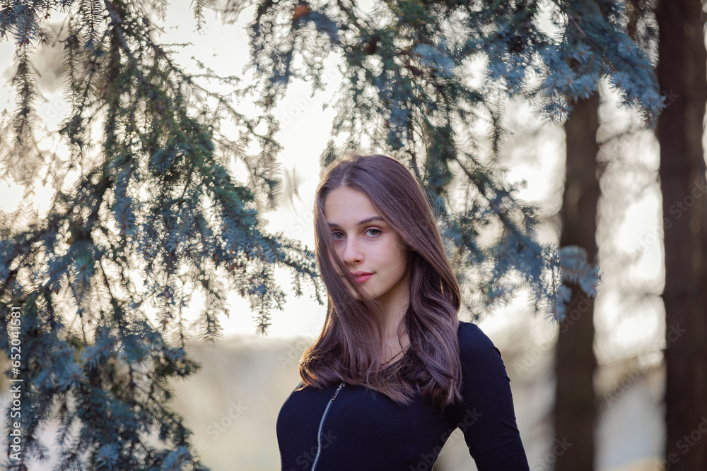 A girl stands near a blue spruce in an autumn park.