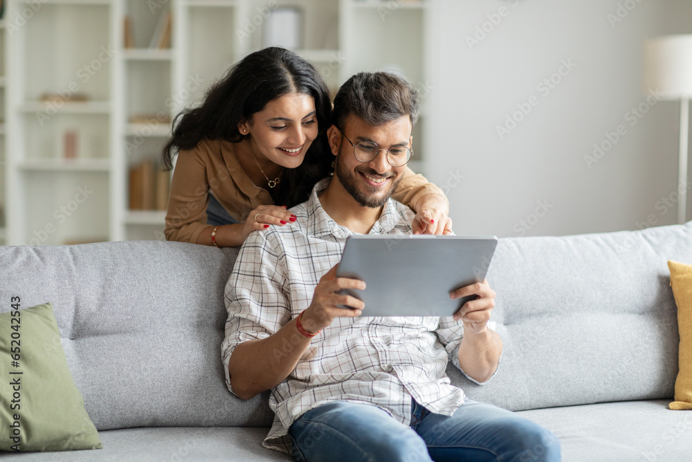 Happy indian man and woman sitting on couch, hugging and using digital tablet, looking at pad screen and smiling, using newest gadget at home interior - obrazy, fototapety, plakaty 