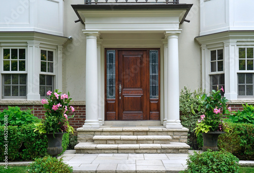 Elegant wood grain front door of home with portico entrance surrounded by flowers and shrubs