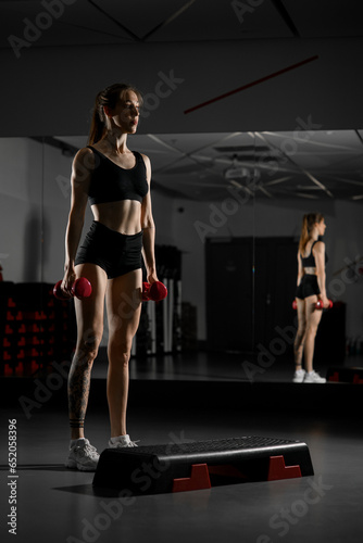 Attractive woman does sports exercises with dumbbells, standing on step platform in gym against mirror background