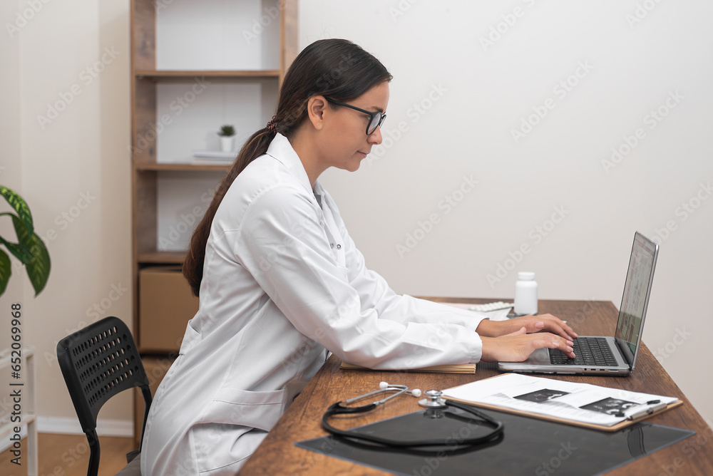 Φωτογραφία Young doctor in glasses enters information of patient diagnosis on laptop side v