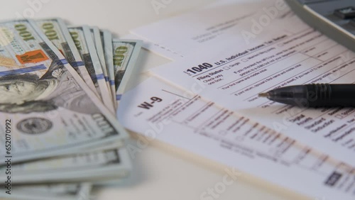 American dollar tax bill, pen calculator and U.S. money. 1040 and W9 individual tax form are placed on desk of American citizen. rotate camera footage photo