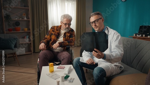 Full-size shot of a doctor checking up the condition of his patient at home. He is talking to an eldery man on a wheelchair, showing a x-ray picture, explaining its meaning.