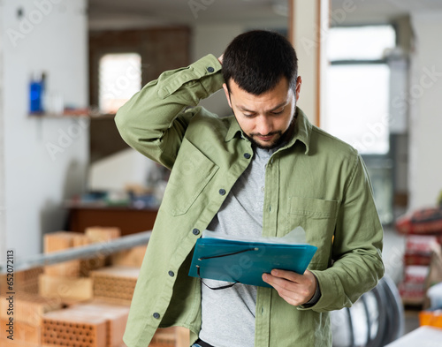 Puzzled and confused young bearded man standing in his house with papers in hands during renovations, determining scope of work