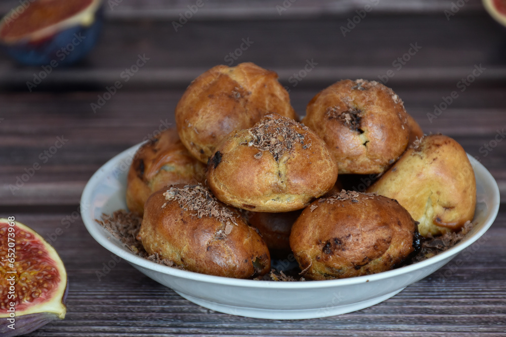 Delicious homemade profiteroles cakes with cocoa cream filling and chocolate chips. Choux pastry, cream puffs on plate, fig halves on wooden table background. Delicious homemade profiteroles cakes with cocoa cream filling and chocolate chips. Choux pastry, cream puffs on plate, fig halves on wooden table background.