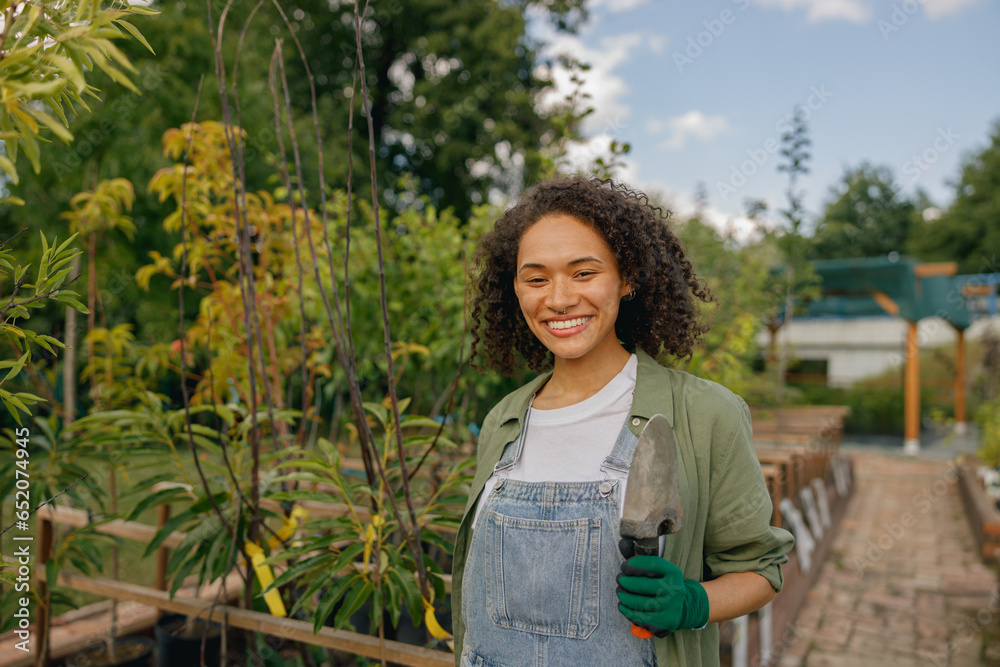 Female gardener standing with hand trowel and looks camera on plants background Female gardener standing with hand trowel and looks camera on plants background