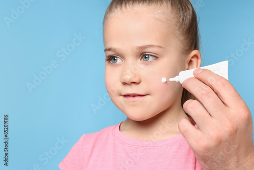 Father applying ointment onto his daughter's cheek on light blue background, space for text