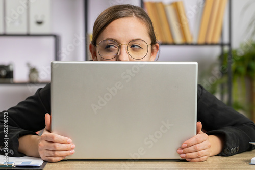 Slika na platnu Confident business woman hiding behind laptop computer, looking at camera, spying his colleagues working, peeping