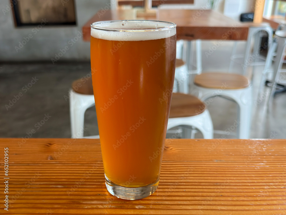 A tall willi becher shaped glass of red ale beer with froth on the top. The liquid alcohol has a vibrant orange tint. The glass is on a wooden table with chairs in the background. 
