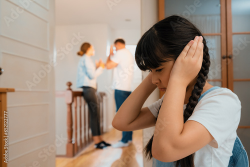 Stressed and unhappy young girl huddle in corner, cover her ears blocking sound of her parent arguing in background. Domestic violence at home and traumatic childhood develop to depression. Synchronos