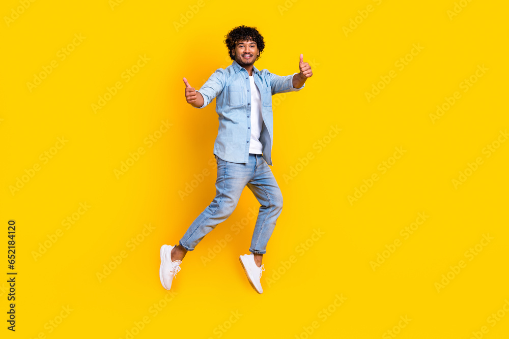 Full length photo of funky cool man wear jeans shirt jumping high thumbs up empty space isolated yellow color background