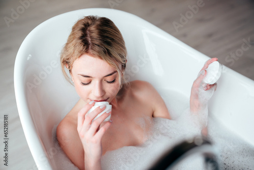 Young beautiful sexy woman having fun while lying in bathtub full of foam at home. Charming smiling model relaxing in luxury bath interior. Female holding cosmetic product, smells jar of cream