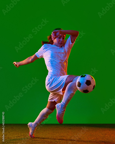 Dynamic image of young woman, football player in motion, kicking ball in jump with knee against green studio background in neon light