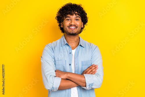 Photo of toothy beaming successful man with curly hairdo dressed denim shirt holding arms crossed isolated on yellow color background