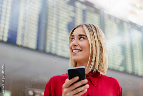 Smiling young businesswoman with cell phone at arrival departure board at the airport photo