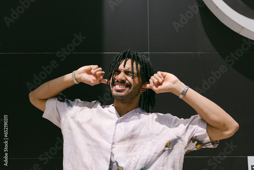Smiling young man with fingers in ears standing near black wall