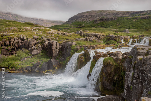 three streams of whitewater form the Baejarfoss waterfall in the Dynjandi park in Iceland with tundra vegetation and a cloudy sky in the background