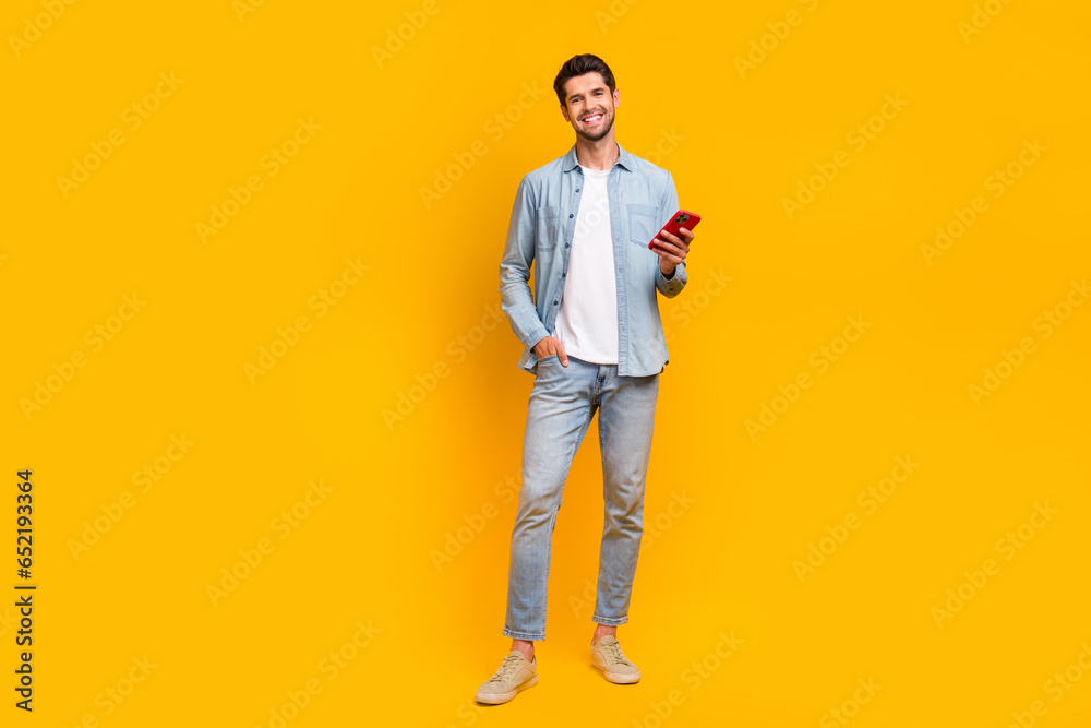 Full length photo of nice positive man wear trendy jeans clothes use modern device iphone samsung isolated on yellow color background Full length photo of nice positive man wear trendy jeans clothes use modern device iphone samsung isolated on yellow color background