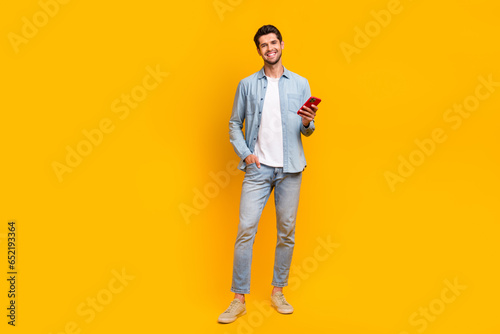 Full length photo of nice positive man wear trendy jeans clothes use modern device iphone samsung isolated on yellow color background