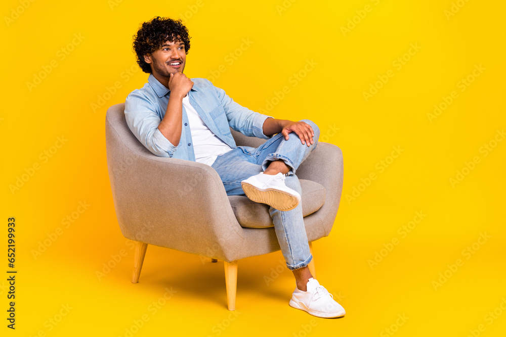 Full length photo of positive minded man wear jeans shirt sit in armchair look empty space finger chin isolated on yellow color background Full length photo of positive minded man wear jeans shirt sit in armchair look empty space finger chin isolated on yellow color background