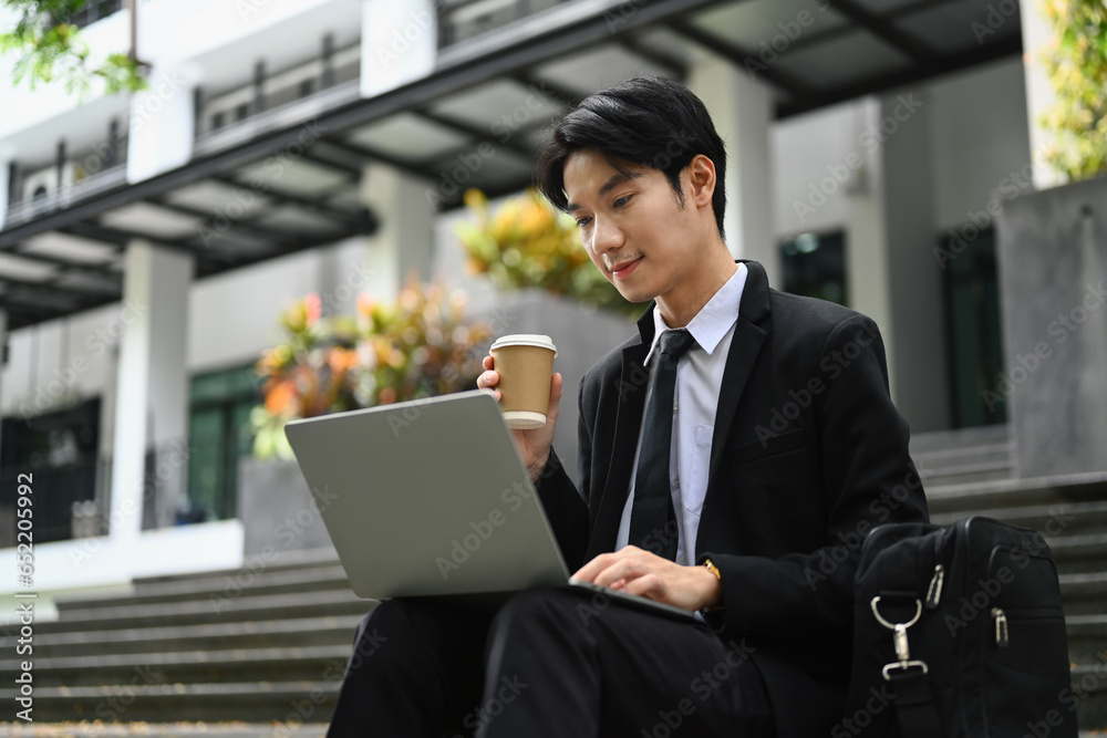 Attractive young businessman sitting on stairs and checking email on laptop before going to the office