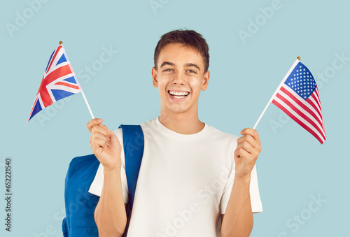 Happy male student learning English language. Studio portrait of school boy with backpack holding American and British flags, looking at camera and smiling isolated on light blue background