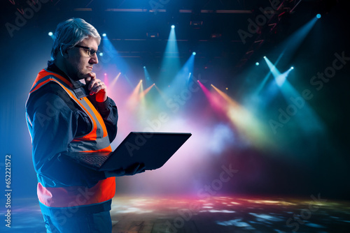 Lighting engineer. Man in hangar with searchlights. Worker sets up equipment in concert hall. Lighting engineer scratches his chin. Gray-haired man master for setting up concert equipment