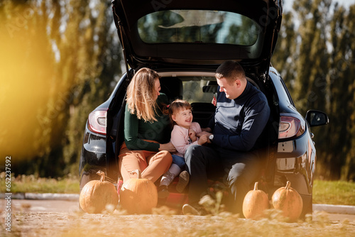 Family day. Happy mom, dad and little child daughter sit at car trunk, look at camera at autumn park with pumpkins, enjoy spend time together at weekend. Parental care and happy carefree childhood